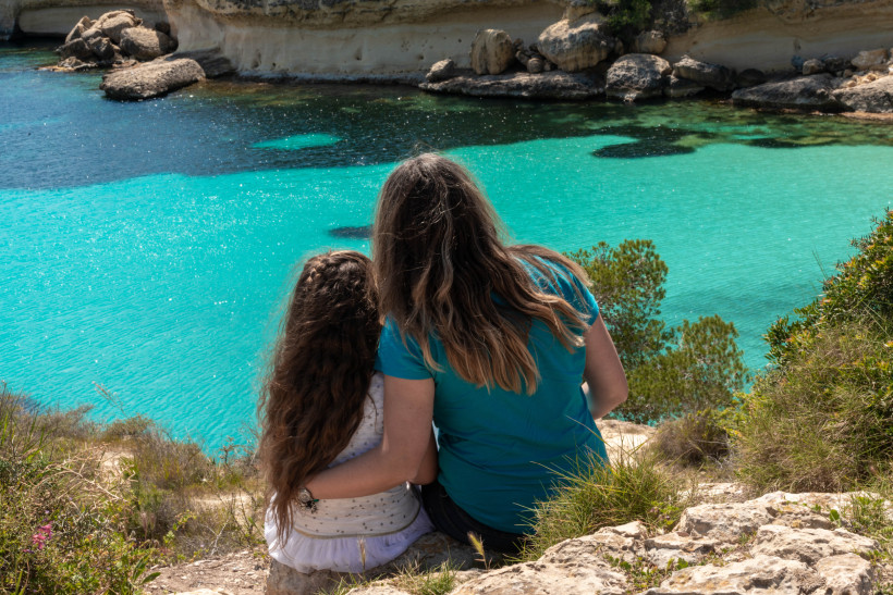 Erwachsener und Kind sitzen an felsiger Küste mit Blick auf ruhiges, türkisfarbenes Wasser