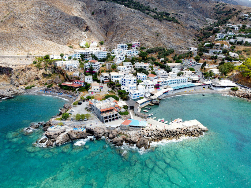 Luftaufnahme des Küstendorfs Loutro auf Kreta mit weißen Häusern am Hang, kleinen Booten im türkisblauen Meer und felsiger Berglandschaft im Hintergrund