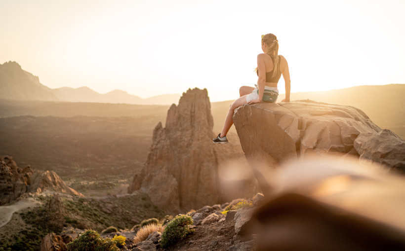 Frau sitzt auf einem Felsen und blickt bei Sonnenuntergang über eine felsige, weite Vulkanlandschaft