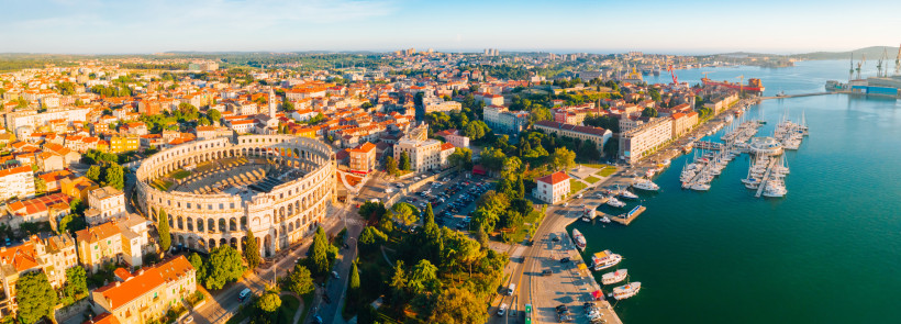 Luftaufnahme der Stadt Pula in Kroatien. Im Vordergrund das gut erhaltene römische Amphitheater, dahinter die Altstadt mit roten Ziegeldächern. Rechts davon ein Jachthafen mit zahlreichen Booten am türkisblauen Meer. Im Hintergrund moderne Stadtviertel un