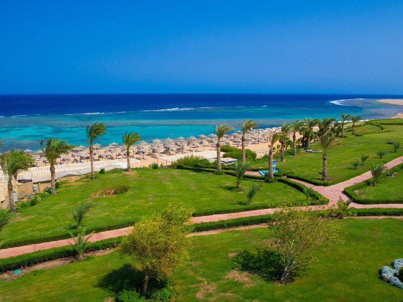 Ägypten - Marsa Alam Grüne Hotelanlage mit Palmen und Wegen, dahinter Sandstrand mit Sonnenschirmen und türkisfarbenem Meer. Der Himmel ist blau und klar.