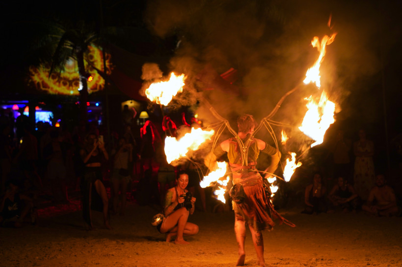Thailand - Koh Samui Das Bild zeigt eine faszinierende Feuershow bei Nacht an einem tropischen Strand. Im Vordergrund steht ein Performer in einem kunstvollen Kostüm mit flügelartigen Gestängen, an denen mehrere Flammen brennen. Um ihn herum tanzen Feuerwirbel, während Rauch