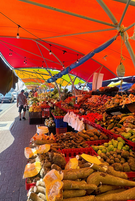 Curaçao Langer Marktstand unter bunten Zeltdächern auf einem Straßenmarkt. Die Auslagen sind prall gefüllt mit tropischen Früchten und Gemüse wie Bananen, Papayas, Yamswurzeln, Kürbis und Avocados. Ein Mann mit Einkaufstasche schlendert entlang der Stände.