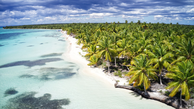 Langer weißer Sandstrand mit seichtem, türkisfarbenem Wasser. Dichter Palmenwald erstreckt sich entlang der Küste, einzelne Menschen spazieren am Strand. Im Hintergrund dramatischer Himmel mit dunklen Wolken über dem hellen Meer.
