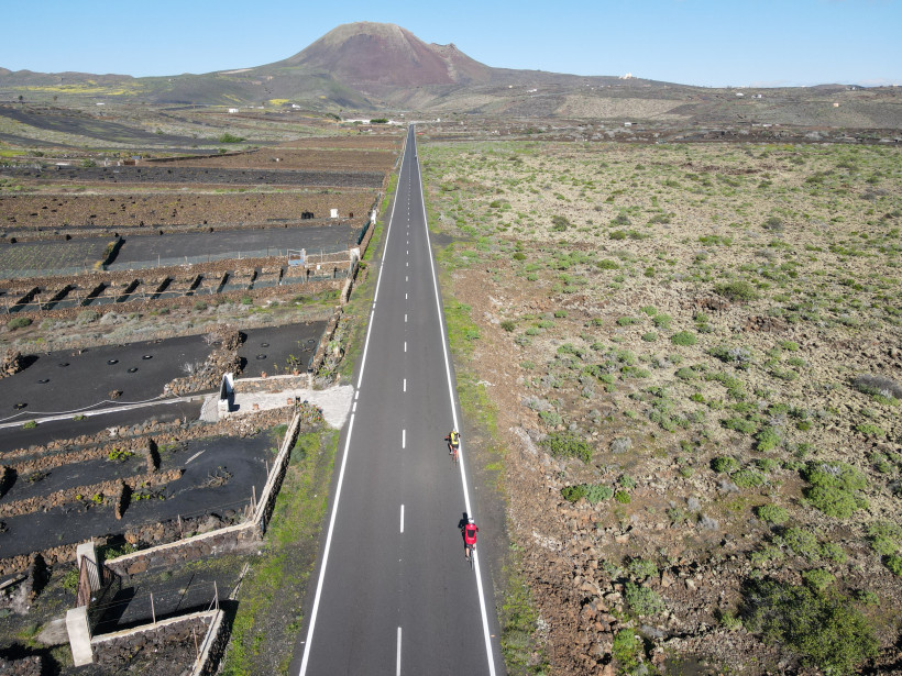 Lanzarote Zwei Radfahrer fahren auf einer langen, geraden Straße durch eine trockene Vulkanlandschaft mit Feldern und niedriger Vegetation. Im Hintergrund erhebt sich ein kegelförmiger Vulkan.