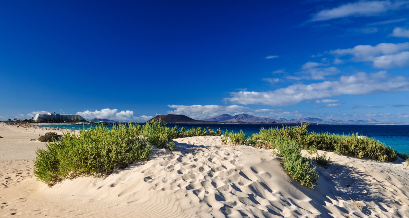 Fuerteventura Blick über eine helle Sanddüne mit grüner Vegetation auf einen Strand mit Hotelgebäuden, türkisblauem Meer und einer Vulkanlandschaft im Hintergrund unter tiefblauem Himmel.