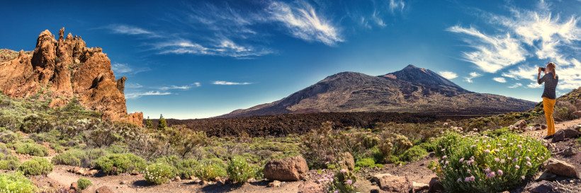 Teide-Nationalpark – Vulkanszenerie Eine Frau steht in einer weiten Vulkanlandschaft im Teide-Nationalpark und fotografiert den Teide, Spaniens höchsten Berg. Vorn wachsen kleine Pflanzen und wilde Blumen, im Hintergrund ragen Lavafelder und braune Vulkankuppen unter blauem Himmel auf.