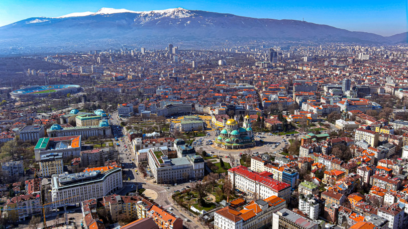 Panoramablick über die Stadt Sofia mit dem Vitosha-Gebirge im Hintergrund. Im Zentrum erkennt man die markante Alexander-Newski-Kathedrale mit ihren goldenen Kuppeln. Die Stadt erstreckt sich weit mit dicht bebauten Wohnvierteln, modernen Hochhäusern und 