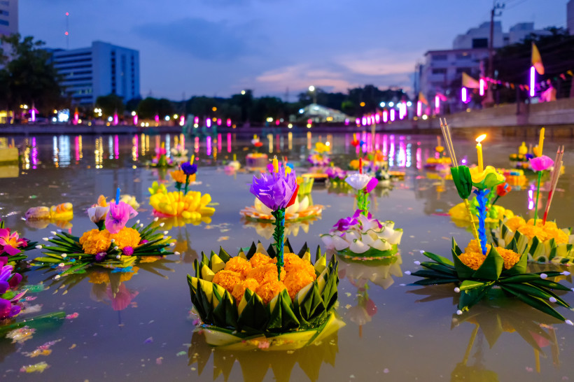 Viele bunte, blumenverzierte Krathongs mit Kerzen und Räucherstäbchen schwimmen auf einem Wasserkanal bei Abenddämmerung. Im Hintergrund leuchten bunte Lichter und Stadthäuser