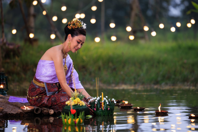 Thailand - Krabi Eine Frau in eleganter thailändischer Kleidung kniet am Ufer eines kleinen Gewässers, um einen mit Blumen, Kerzen und Räucherstäbchen geschmückten Krathong behutsam aufs Wasser zu setzen. Die Szene spielt bei Nacht, im Hintergrund sind warme Lichtpunkte d