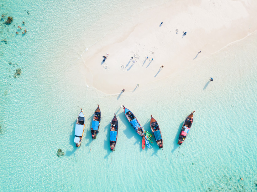Thailand - Phuket Drohnenaufnahme von sieben traditionellen Longtail-Booten, die nebeneinander im türkisfarbenen, klaren Wasser einer Sandbank auf Phuket liegen. Einige Menschen spazieren barfuß über den hellen Sand.