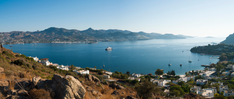 Weitläufiger Blick über eine mediterrane Bucht mit ruhigem Meer, weißen Häusern an der Küste und bergiger Landschaft im Hintergrund