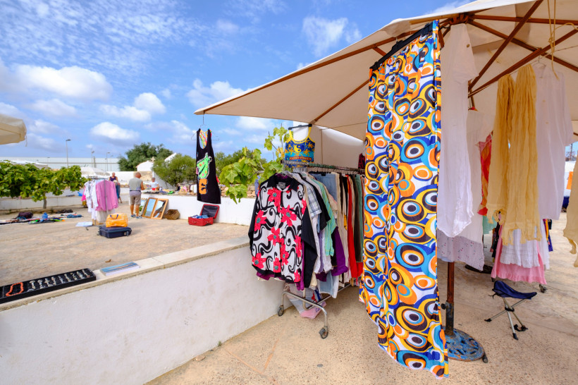 Formentera Offener Marktstand unter einem großen Sonnenschirm, an dem bunte Sommerkleidung auf Kleiderständern hängt. Im Vordergrund auffällige Muster in Orange, Blau und Weiß. Im Hintergrund weitere Stände und Besucher.