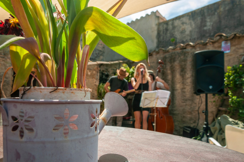 Formentera Live-Musikszene in einem sonnigen Innenhof mit alten Mauern. Im Vordergrund eine Gießkanne mit Pflanzen, im Hintergrund eine Sängerin mit Mikrofon, begleitet von Musikern mit Gitarre und Kontrabass