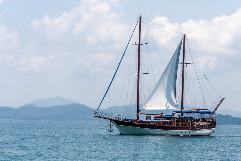 Thailand - Phuket Nahaufnahme eines großen, klassischen Segelboots mit weißen Segeln und Holzdeck, das auf ruhigem, blauen Meer vor den grünen Hügeln von Phuket unterwegs ist. Der Himmel ist leicht bewölkt, die Szene strahlt Ruhe und Weite aus.