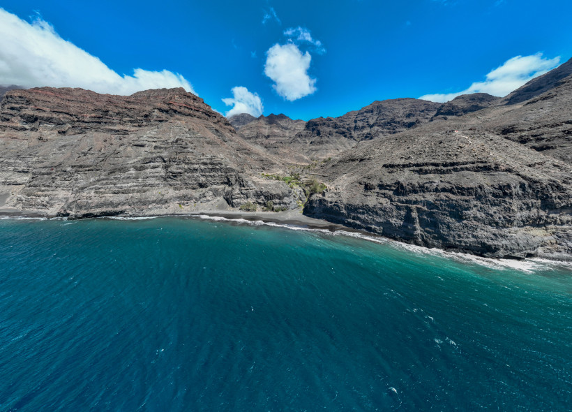 Playa de Güigüí – spektakuläre Luftaufnahme des Naturstrands Ein Luftbild zeigt den Playa de Güigüí an der Westküste Gran Canarias. Der dunkle Sandstrand liegt einsam zwischen steilen, felsigen Berghängen. Das Meer leuchtet tiefblau, kleine Wellen treffen auf den Strand.