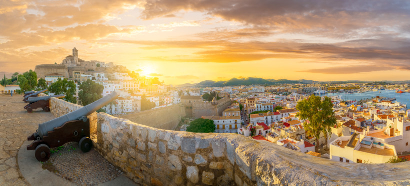 Ibiza Panoramablick auf Ibiza-Stadt bei Sonnenuntergang. Im Vordergrund stehen mehrere alte Kanonen auf der Stadtmauer, dahinter reihen sich weiße Häuser und historische Gebäude aneinander. Am Horizont leuchtet die untergehende Sonne zwischen sanften Hügeln.