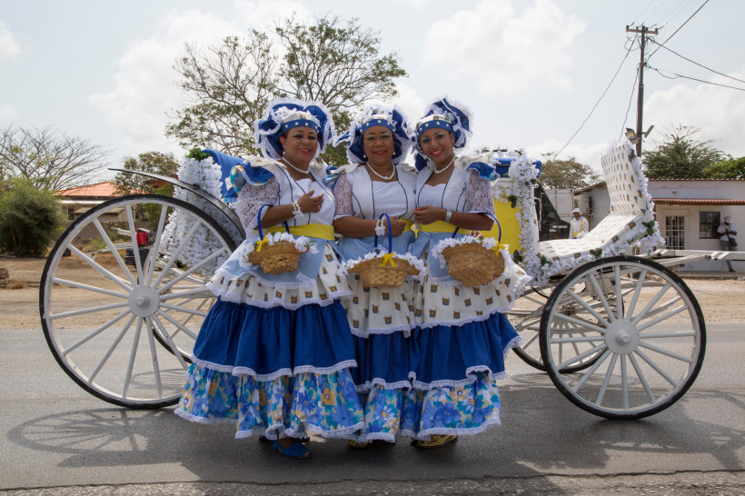 Curaçao Drei Frauen in farbenfroher traditioneller Kleidung stehen nebeneinander vor einer dekorierten weißen Kutsche mit großen Rädern. Sie tragen blau-weiße Kleider mit Spitzenapplikationen, gelben Schärpen und passenden Hauben. Jede hält ein kleines Körbchen i