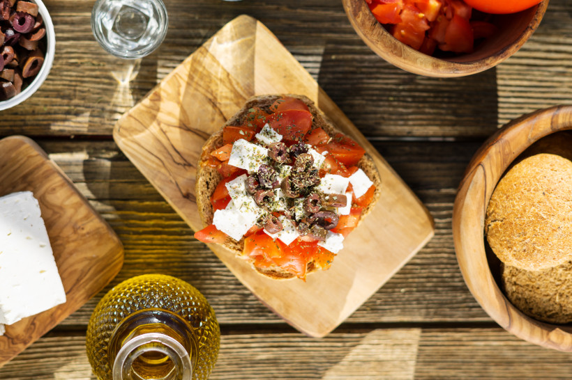 Kreta Ein traditioneller griechischer Dakos auf einem Holzbrett: Geröstetes Gerstenbrot belegt mit gewürfelten Tomaten, Fetakäse, Oliven und Kräutern. Umgeben von weiteren Zutaten wie Oliven, Tomaten, Brot, Feta und einer Flasche Olivenöl auf einem rustikalen H