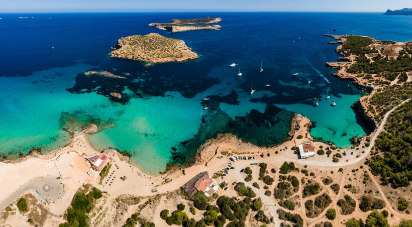 Luftaufnahme der Bucht Cala Conta mit türkisfarbenem Wasser, vorgelagerten Inseln, Strandabschnitt und Segelbooten