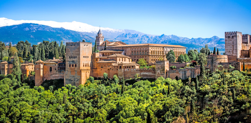 Die Alhambra in Granada mit ihren rötlich-braunen Türmen vor schneebedeckten Bergen der Sierra Nevada, umgeben von grünen Bäumen.