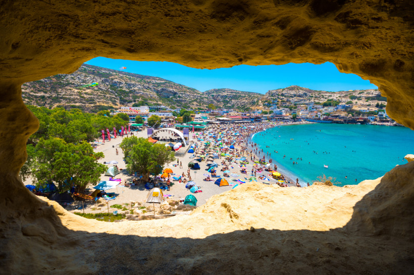 Kreta Blick aus einer natürlichen Felsengrotte auf den belebten Strand von Matala, Kreta. Der Sandstrand ist voller bunter Sonnenschirme, Badetücher und Menschen. Am Rand befindet sich eine Bühne mit Festivalcharakter. Dahinter erstreckt sich eine Bucht mit tür