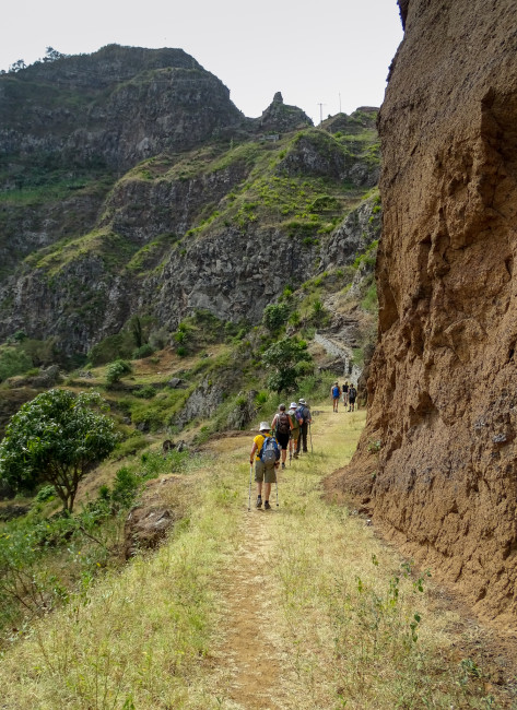 Wandergruppe mit Rucksäcken und Wanderstöcken auf einem schmalen Pfad entlang einer Felswand in einer bergigen, grün bewachsenen Landschaft. Die Wandernden bewegen sich durch eine tiefe Schlucht, umgeben von steilen Hängen.