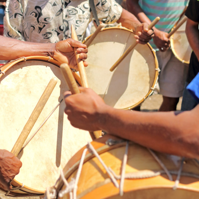 Mehrere Menschen spielen traditionelle Trommeln mit Holzstöcken bei einem Straßenfest. Die Szene zeigt Nahaufnahmen von Händen, Trommeln und rhythmischer Bewegung. Die Kleidung ist sommerlich und farbenfroh.