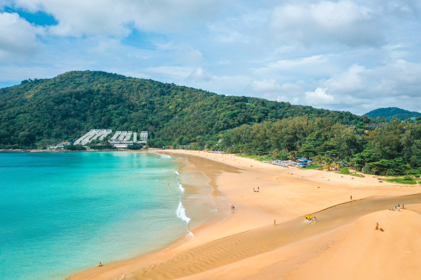 Blick auf den Nai Harn Beach mit breitem Sandstrand, türkisfarbenem Meer und bewaldeten Hügeln im Hintergrund