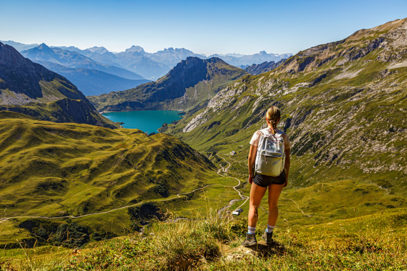 Österreich Panoramablick über grüne Alpentäler mit türkisblauem Bergsee; Wanderin mit Rucksack steht am Grat bei klarem Sommerwetter.