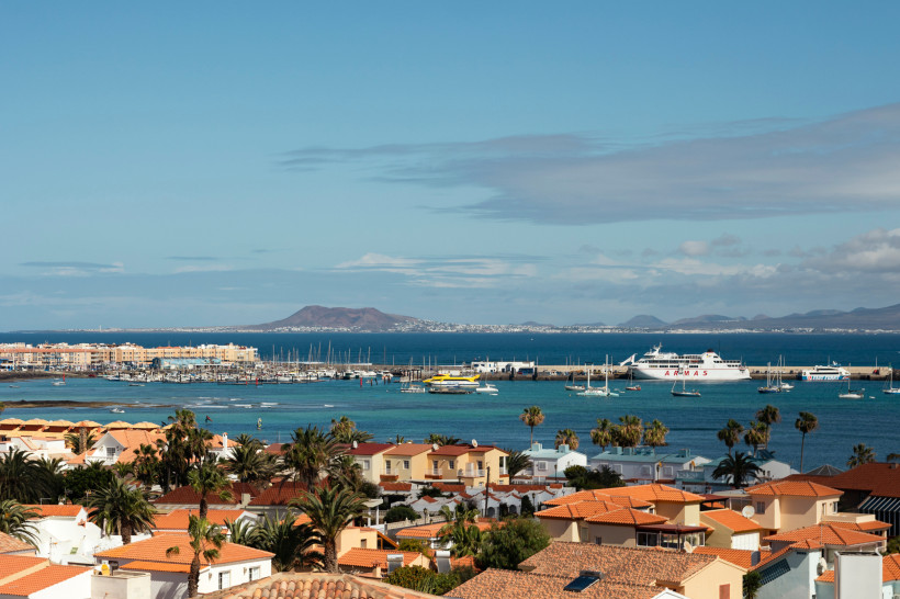 Blick über Corralejo auf Fuerteventura: Hafen mit Fähren und Yachten, türkisfarbenes Meer, rotgedeckte Dächer und Lanzarote am Horizont.