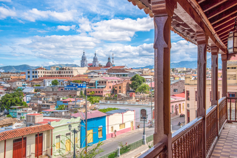 Panoramablick von einem Holzbalkon auf die farbenfrohen Häuser der Altstadt von Santiago de Cuba mit der imposanten Kathedrale im Hintergrund und den Bergen der Sierra Maestra.