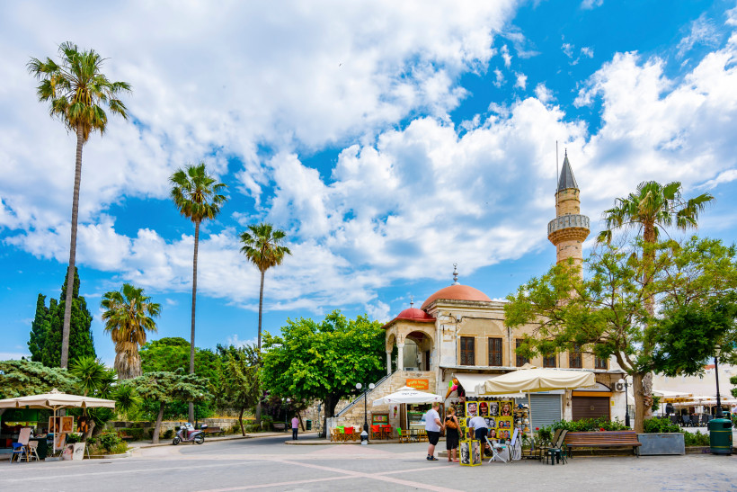 Kos-Stadt Lebhafter Platz in der Stadt Kos mit Palmen, einem Verkaufsstand und dem historischen Gebäude der Defterdar-Moschee mit roter Kuppel und Minarett. Menschen bummeln gemütlich unter blauem Himmel mit dekorativen Wolken.