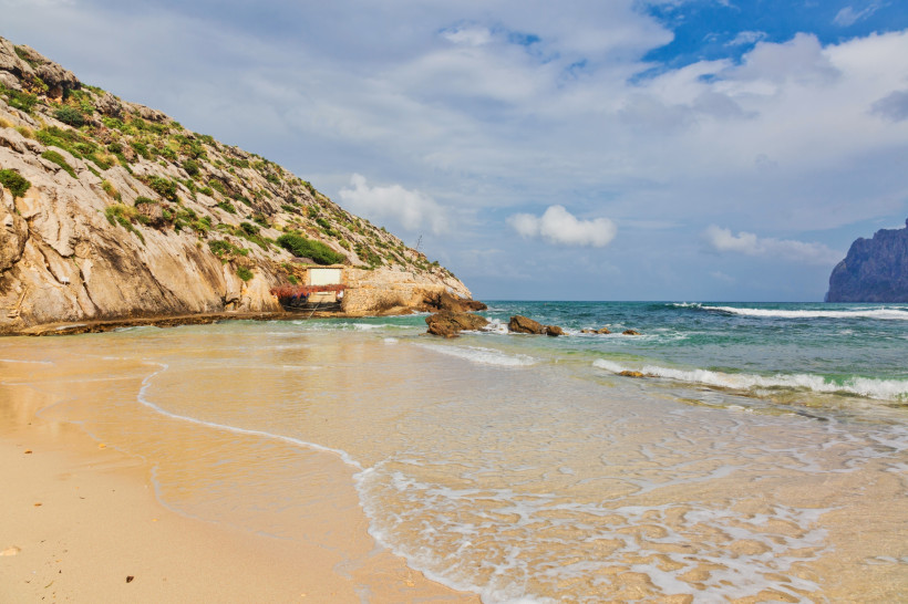 Sandstrand der Cala Barques in Cala San Vicente mit flachem Ufer, Felswand und Blick auf das offene Meer.