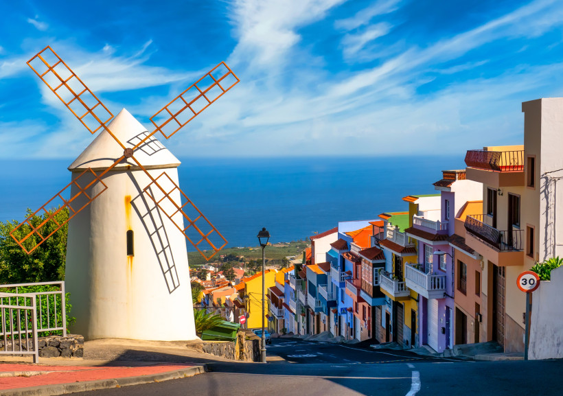 Eine weiße traditionelle Windmühle steht an einer steilen Straße mit bunten Häusern in Gelb, Blau und Orange. Im Hintergrund der blaue Atlantik unter einem hellen Himmel mit leichten Wolkenstreifen.