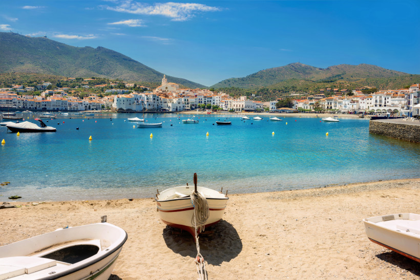 Blick von einem Sandstrand mit kleinen Booten auf das klare blaue Meer und die malerische Küstenstadt Cadaqués in Katalonien, Spanien. Im Hintergrund steigen weiße Häuser an einem Hügel auf, überragt von einer Kirche mit hellem Turm.