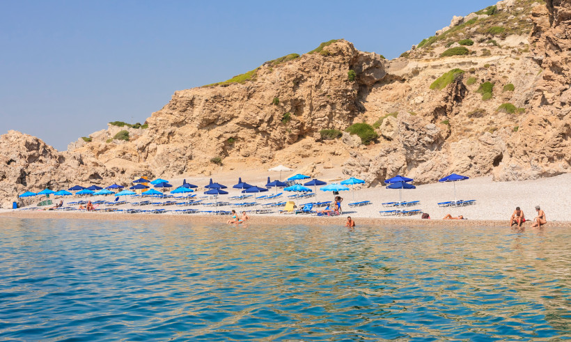 Felsige Badebucht auf Rhodos mit türkisfarbenem Meer, Strand mit blauen Sonnenschirmen und Liegen sowie kleinen Booten im Wasser