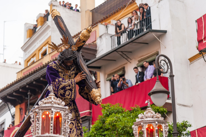 Fuerteventura Prunkvolle Christusfigur in violett-goldenem Gewand trägt ein Kreuz während einer Prozession durch eine Straße mit Zuschauern auf Balkonen. Die Szene ist geschmückt mit silbernen Leuchtern und rotem Stoff