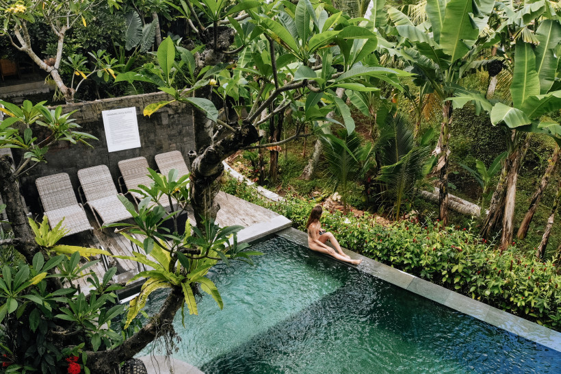 Bali Eine Frau sitzt entspannt am Rand eines privaten Pools inmitten tropischer Vegetation auf Bali. Umgeben von Palmen, Bananenpflanzen und dichtem Grün wirkt der Poolbereich ruhig und abgeschieden. Neben dem Pool stehen mehrere leere Sonnenliegen auf einer H