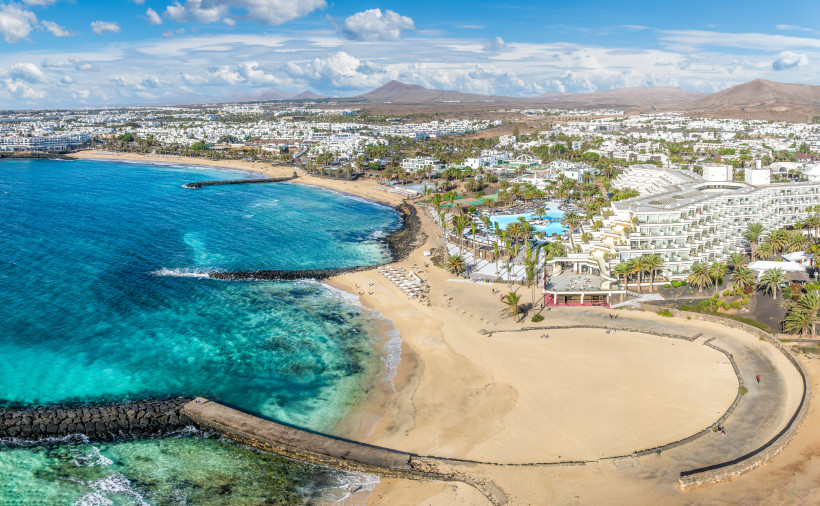 Lanzarote Panoramaaufnahme eines Ferienorts mit weißen Gebäuden, Sandstrand und türkisblauem Meer. Wellenbrecher schützen die Buchten, im Hintergrund liegen Hügel unter blauem Himmel.