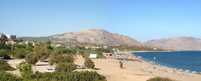 Panoramablick auf einen Sandstrand auf Rhodos mit Badegästen, Sonnenschirmen und einer Berglandschaft im Hintergrund an der griechischen Küste.