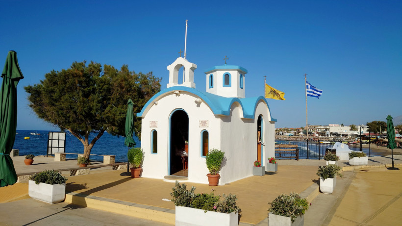Kleine weiß-blaue Kapelle am Hafen von Analipsi auf Kreta, im Hintergrund Meer und Flaggen