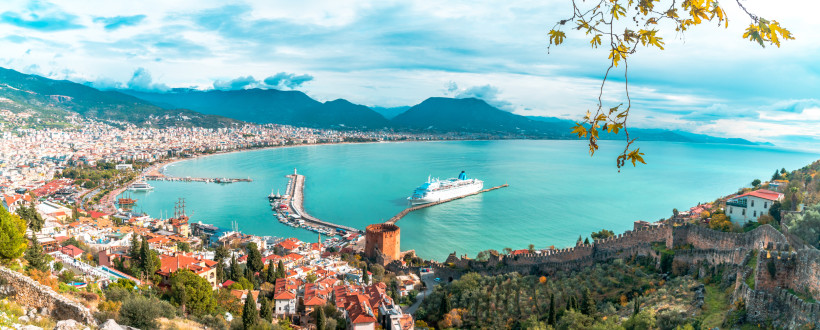Das Bild zeigt Alanya an der türkischen Riviera mit der historischen Altstadt, dem Roten Turm und der Stadtmauer im Vordergrund. Eine weite Bucht, ein Kreuzfahrtschiff im Hafen und das türkisfarbene Meer treffen auf die moderne Stadt und grüne Berge im Hi