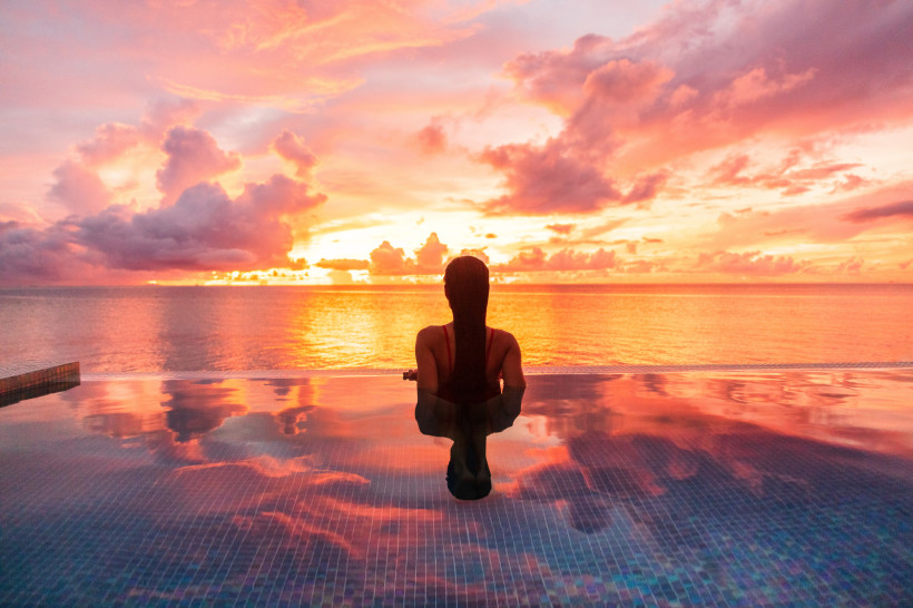 Jamaika Eine Frau sitzt mit dem Rücken zur Kamera in einem Infinity-Pool und blickt auf das offene Meer. Der Himmel ist in dramatischem Orange, Rosa und Violett gefärbt, da die Sonne gerade untergeht. Die Farben spiegeln sich eindrucksvoll im ruhigen Wasser des P