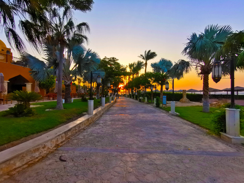 Ägypten - Makadi Bay Sonnenuntergang auf einer palmengesäumten Promenade einer Hotelanlage, mit Blick auf das Meer. Rechts und links sind Laternen, Palmen und mediterrane Gebäude zu sehen.