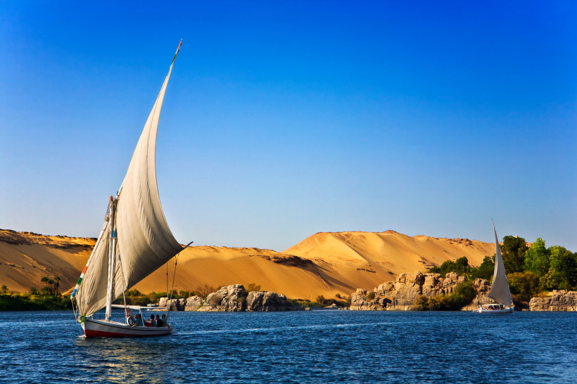 Ägypten Traditionelles ägyptisches Segelboot, eine Feluke, gleitet über den Nil vor einer Landschaft aus goldenen Sanddünen und Felsen. Das Segel ist hoch aufgezogen, der Himmel klar und blau.