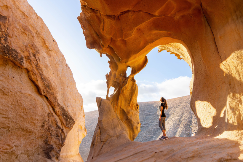 Fuerteventura Frau steht zwischen bizarren, orangefarbenen Felsformationen und blickt staunend nach oben. Im Hintergrund karge Hügel unter blauem Himmel mit Wolken