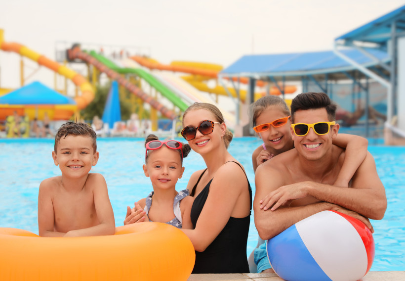 Ausflug einer Familie in einen Wasserpark am Pool mit Wasserrutschen Familie im Wasserpark am Beckenrand mit Schwimmreifen und Wasserball, im Hintergrund Wasserrutschen