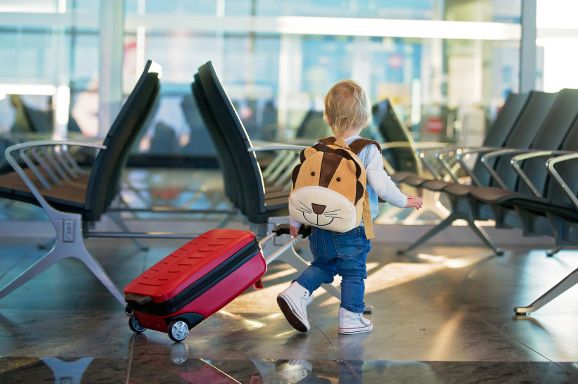 Kleines Kind am Flughafen mit Rucksack und rotem Rollkoffer im Wartebereich