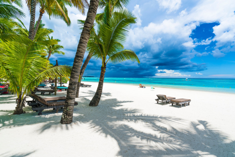 Weißer Sandstrand mit Palmen und Sonnenliegen auf Mauritius, türkisblaues Meer und dramatische Wolken am Himmel